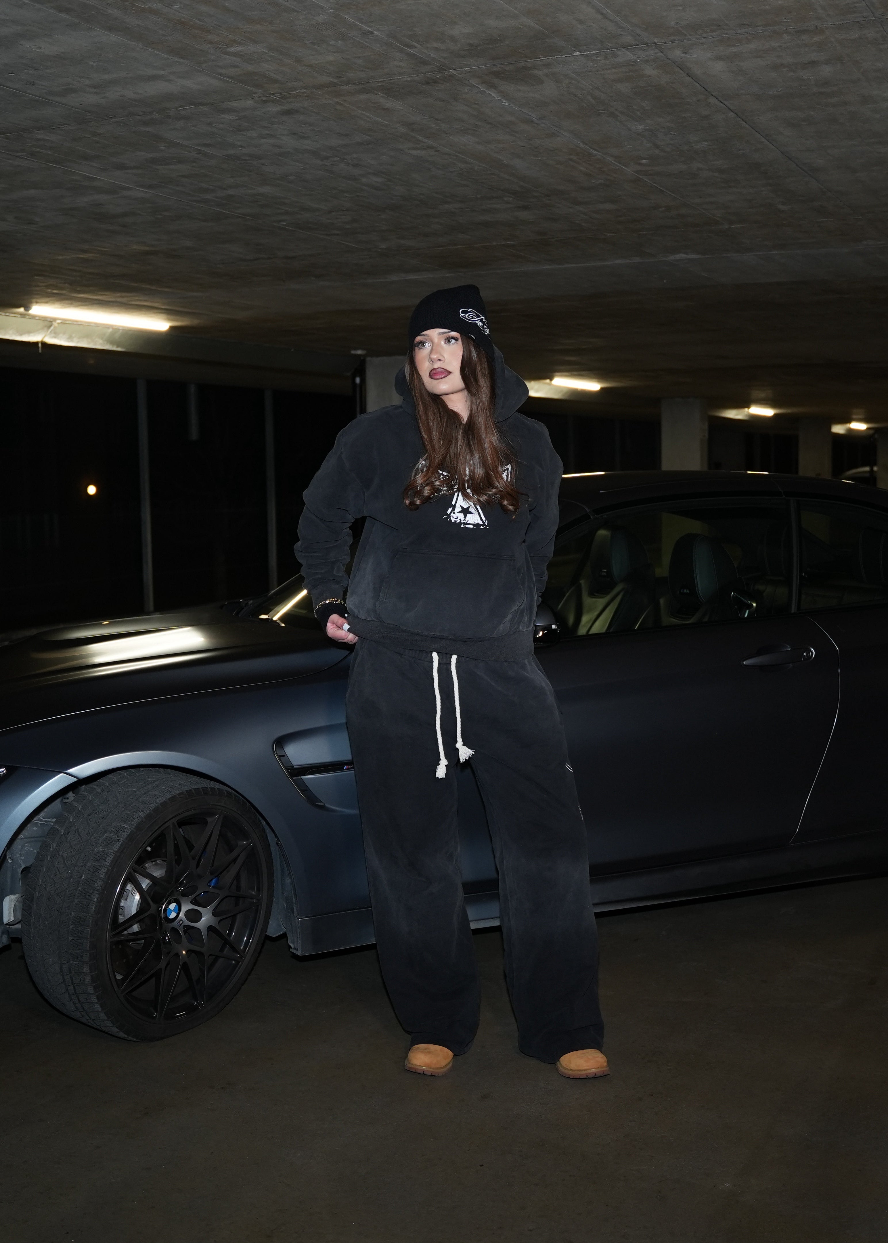 Person wearing a black hoodie branded "evoke" and pants standing next to a car in a dark parking garage.
